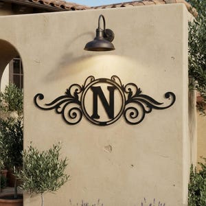 May include: A black metal wall sign featuring a central circular frame enclosing the capital letter N, flanked by symmetrical scrollwork and decorative flourishes. The piece is mounted on a light-coloured stucco wall beneath a dark metal light fixture.