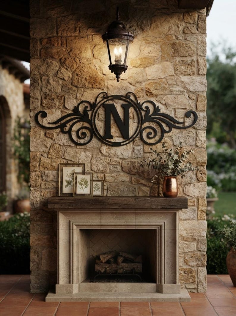 May include: A black metal wall sign featuring a central letter N surrounded by decorative scrollwork. The sign is mounted on a stone wall above a fireplace mantel, which holds three framed botanical prints and a copper vase with greenery.