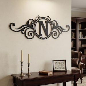 May include: A black metal wall art piece featuring a central circular frame enclosing the capital letter N, flanked by symmetrical scrollwork and leaf-like flourishes. The decorative sign is mounted horizontally on a light-coloured wall.