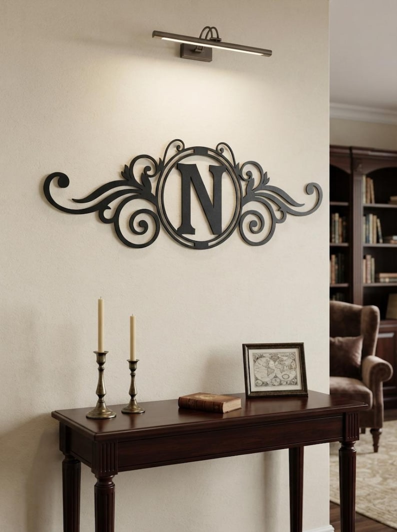 May include: A black metal wall art piece featuring a central circular frame enclosing the capital letter N, flanked by symmetrical scrollwork and leaf-like flourishes. The decorative sign is mounted horizontally on a light-coloured wall.