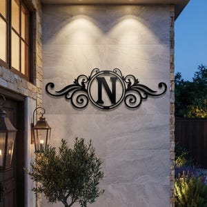 May include: A black metal wall sign featuring a central circular frame enclosing the capital letter N, flanked by symmetrical decorative scrollwork and flourishes. The sign is mounted on a light-coloured stone wall surface with recessed lighting.