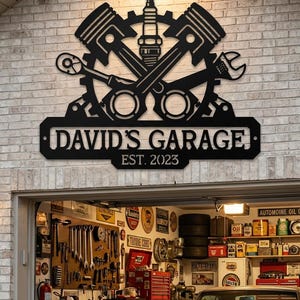 Könnte beinhalten: Ein schwarzes Metall-Wandschild mit einem Zahnrad, zwei gekreuzten Kolben, einer Zündkerze, einer Ratsche und einem verstellbaren Schraubenschlüssel. Das Schild zeigt den Text DAVID'S GARAGE und EST. 2023 in fetter Schrift mit Befestigungslöchern.