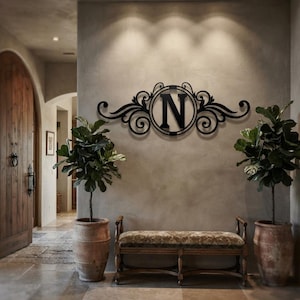 May include: A black metal wall sign featuring a central circular frame enclosing the capital letter N, flanked by symmetrical decorative scrollwork and flourishes. The piece is mounted on a wall above a bench, displaying a dark finish.