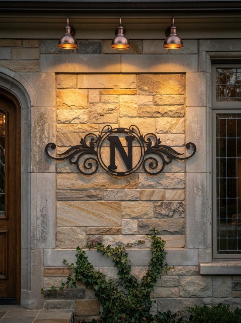 May include: A black metal wall sign featuring a central circular frame enclosing the capital letter N, accented with decorative scrollwork on both sides. The sign is mounted on a light-coloured stone wall beneath three downward-facing light fixtures.