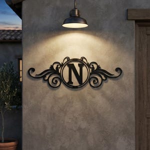 May include: A black metal wall sign featuring a central circular frame containing the capital letter N, flanked by symmetrical scrollwork designs. The sign is mounted on a textured grey wall directly beneath a downward-facing metal light fixture.