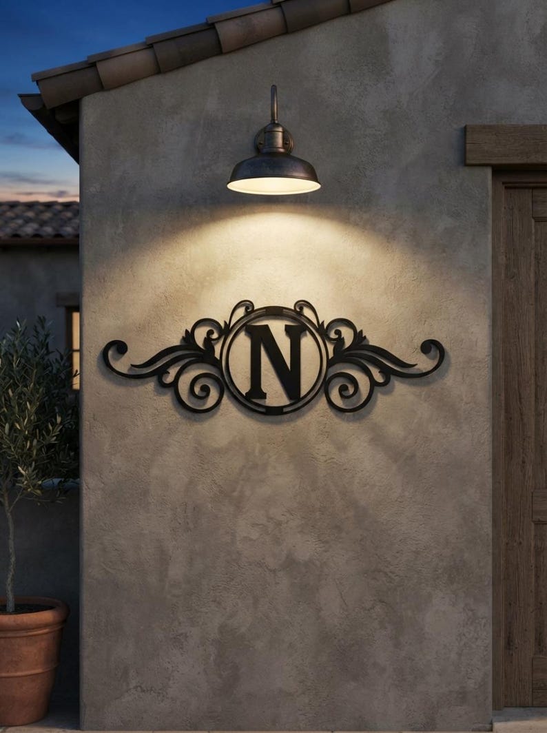 May include: A black metal wall sign featuring a central circular frame containing the capital letter N, flanked by symmetrical scrollwork designs. The sign is mounted on a textured grey wall directly beneath a downward-facing metal light fixture.
