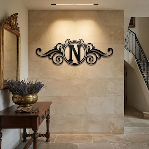 May include: A black metal wall sign featuring a central circular frame containing the capital letter N, flanked by symmetrical decorative scrollwork and flourishes. The piece is mounted on a light-coloured stone wall above a wooden console table.