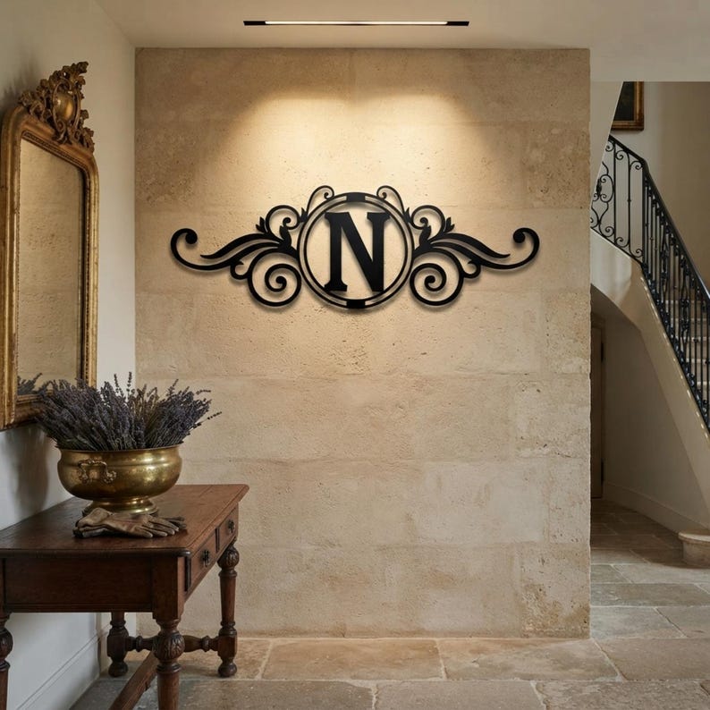 May include: A black metal wall sign featuring a central circular frame containing the capital letter N, flanked by symmetrical decorative scrollwork and flourishes. The piece is mounted on a light-coloured stone wall above a wooden console table.