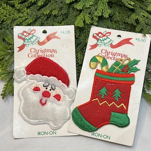 May include: Two iron-on embroidered patches on white cardboard backing cards labeled Christmas Collection. One patch is a Santa Claus face with a red hat and white beard. The other is a red Christmas stocking with green trim, two small green trees, a candy cane, and a gift.