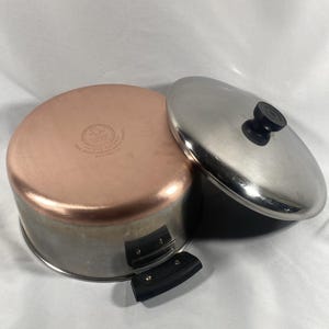 May include: A copper-bottomed saucepan with a stainless steel lid. The pan has a silver exterior and a copper interior. The lid has a black handle. The pan's handle is black. The pan is designed for cooking.