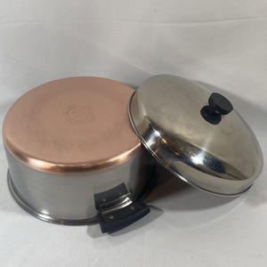 May include: A stainless steel cooking pot with a copper-colored base and a matching stainless steel lid. The pot features a black handle attached to the side, and the lid has a central black knob. The base shows a circular stamped logo.