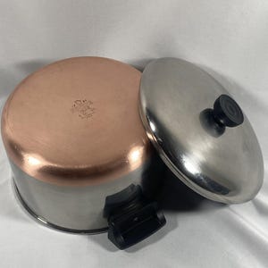 May include: A stainless steel and copper-bottomed saucepan with a lid. The copper bottom is visible, along with a black handle and a stainless steel lid with a black knob. The pan is designed for cooking.