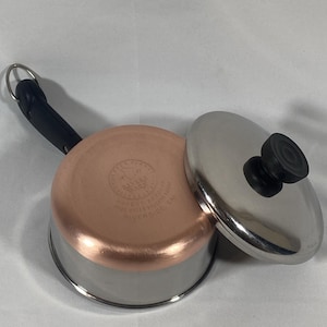 May include: A Revere Ware saucepan with a copper-clad bottom and a stainless steel body. The pan has a black handle and a stainless steel lid with a black knob. The pan is approximately 6 inches in diameter.
