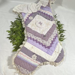 May include: A quilted Christmas stocking in shades of lavender, purple, and white. The stocking features a patchwork design with floral patterns, lace trim, a ruffled top edge, and a central diamond-shaped panel with embroidered floral details.