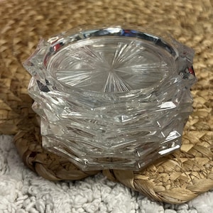 May include: A stack of clear glass coasters featuring a faceted, geometric edge design and a central sunburst pattern radiating from the middle. The coasters are transparent with a reflective surface, showcasing a multi-layered, circular structure.