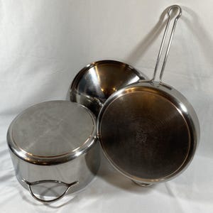 May include: A set of stainless steel cookware including a round pot with two side handles and a skillet with a long wire handle. Both pieces feature a polished metallic finish and a circular stamp on the bottom surface, showcasing a classic kitchen design.