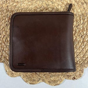 May include: A square, dark brown leather wallet featuring a smooth finish, rounded corners, and a zipper closure along the top edge. A small, rectangular metal plaque with the embossed brand name is attached to the lower left corner of the front.
