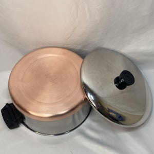 May include: A stainless steel cooking pot with a copper-colored base and a black handle, accompanied by a matching stainless steel lid featuring a black knob. The pot and lid are positioned side-by-side, showcasing their metallic finish.