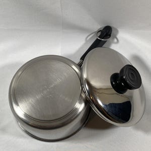May include: A stainless steel saucepan with a lid. The saucepan has a long black handle and a lid with a black knob. The pot and lid are made of shiny, brushed metal. The lid is slightly ajar, revealing the interior of the pot.