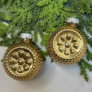 May include: Two round gold-colored glass ornaments featuring a raised floral pattern on the front and a textured dimpled surface on the sides. Each ornament is topped with a silver-colored metal cap for hanging, reflecting a metallic finish.