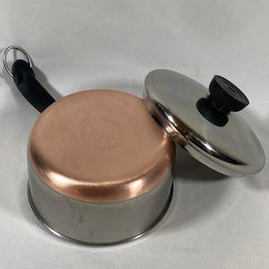 May include: A stainless steel and copper saucepan with a lid. The pot has a copper bottom and a stainless steel body. The lid is stainless steel with a black handle. The handle on the pot is black and has a metal loop.