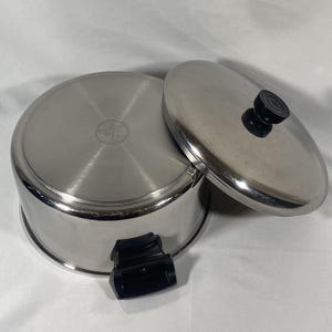 May include: Stainless steel saucepan with lid. The pot is silver with a black handle and a matching lid with a black knob. The lid is slightly ajar, revealing the interior of the pot. The pot is sitting on a white surface.