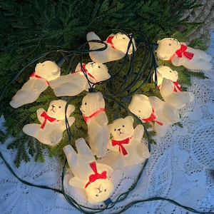 May include: A string of decorative lights featuring multiple white plastic teddy bear figures. Each bear is moulded with a textured surface and wears a bright red painted bow around its neck. The bears are connected by a dark green electrical wire.