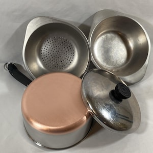 May include: A set of stainless steel cookware including a colander, a deep bowl, and a saucepan with a copper-colored base, a black handle, and a matching lid with a black knob. The pieces feature smooth metallic surfaces and functional designs.