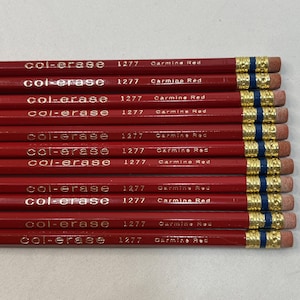 May include: A stack of twelve red Col-Erase 1277 Carmine Red pencils. Each pencil features gold lettering, a gold-colored metal ferrule, a thin blue band, and a pink eraser at the end. The pencils are arranged horizontally in a parallel row.