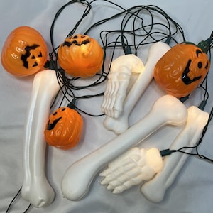 May include: A string of Halloween lights featuring orange plastic jack-o'-lanterns with black facial features, white plastic bones, and white plastic skeletal hands. The decorative light set is connected by a thin black electrical wire.