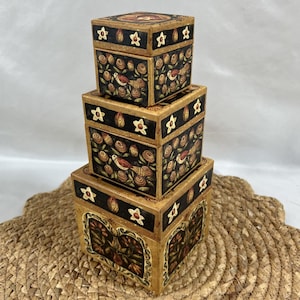 May include: A set of three square nesting boxes stacked vertically. Each box features a black background with hand-painted motifs of birds, swirling floral patterns, and cream-colored star shapes. The edges are finished in a distressed tan color.