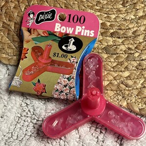 May include: A package of 100 Pixie Bow Pins featuring a pink, three-pronged plastic container filled with small, clear plastic pins. The cardboard header displays the text 100 Bow Pins, a graphic of the pin, and a vintage illustration of a person.