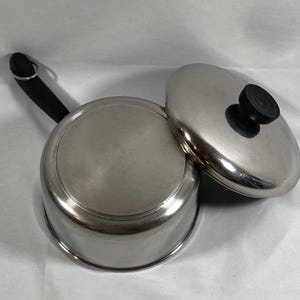 May include: A stainless steel saucepan with a black handle and a matching lid. The lid has a black knob. The pan is sitting on a white surface.