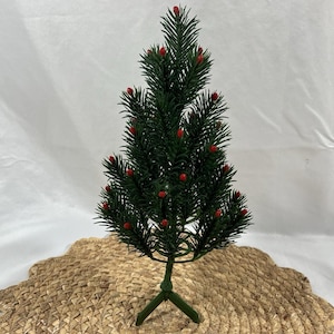 May include: A small artificial Christmas tree with dark green pine needles and numerous tiny red berries scattered throughout the branches. The tree is supported by a simple green three-legged base, creating a compact and upright structure.