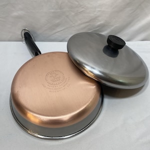May include: A round stainless steel frying pan with a copper-clad base and a black handle. The base features the stamped text: 1801 REVERE WARE STAINLESS STEEL, PATENT 2272609, MADE UNDER PROCESS PATENT 23639, RIVERSIDE, CAL. Includes a matching lid.