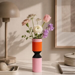May include: A cylindrical vase with a vertical ribbed texture, featuring a color-blocked design of light pink, bright orange, and black sections. The vase holds a bouquet of mixed flowers including a white ranunculus, a pink tulip, and purple sweet peas.