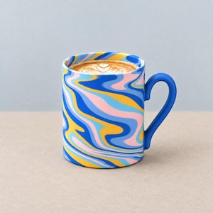 May include: A ceramic mug with a matte finish featuring a swirling marbled pattern of blue, light blue, yellow, and pink. The mug has a solid blue handle and is filled with coffee topped with a white heart-shaped latte art design.