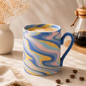 May include: A ceramic mug featuring a swirling marbled pattern in shades of blue, yellow, and soft pink. The mug has a curved handle and is filled with coffee topped with a heart-shaped latte art design. The exterior surface is smooth and glossy.