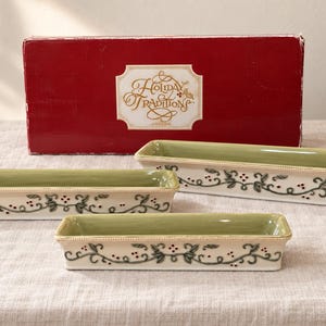 May include: Three rectangular ceramic serving dishes with a light green interior and a cream exterior featuring a dark green vine and red berry pattern. The dishes are positioned in front of a red box labeled Holiday Traditions in gold script.