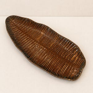May include: An elongated, leaf-shaped ceramic tray with a glossy, dark brown glaze. The surface features a textured pattern of parallel ridges radiating from a central spine, mimicking the natural veins and structure of a large tropical leaf.