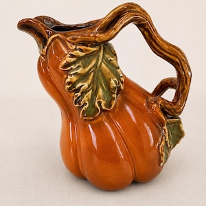 May include: A glossy ceramic pitcher shaped like a pumpkin with a vibrant orange body and a dark brown handle resembling a twisted vine. The pitcher features raised green leaf details on the side and a spout for pouring, with a smooth finish.