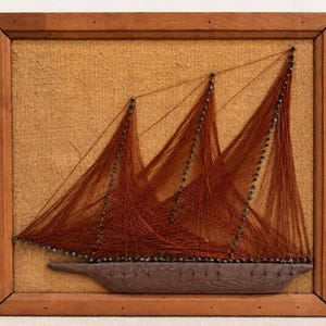 May include: A string art wall hanging featuring a dark brown wooden ship hull mounted on a textured tan burlap background. The sails are created with thin, dark brown thread wrapped around small metal nails, all enclosed in a simple wooden frame.