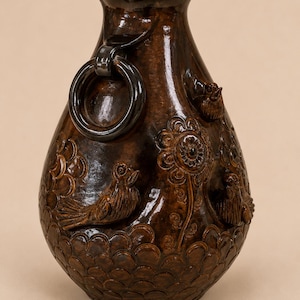 May include: A dark brown glazed ceramic vase featuring a relief pattern of birds, a central flower, and a scalloped base design. A prominent dark metal ring handle is attached to the upper neck of the vessel, which has a smooth, glossy finish.