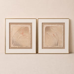 May include: Two framed wall art pieces featuring embossed floral designs on textured, deckled-edge paper. The left print depicts a Bird of Paradise, and the right print shows a Calla Lily. Both are set in gold frames with cream-colored mats.