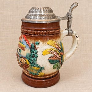 May include: A ceramic beer stein featuring a brown base and rim with a central cream-colored body. The exterior displays a raised, colorful relief of a house, foliage, and wheat stalks. It includes a hinged metal lid and a curved handle.