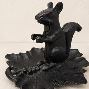 May include: A black cast iron nutcracker shaped like a squirrel standing on a base designed as a large leaf with a cluster of grapes. The squirrel is positioned upright with its tail curved upward, and the entire object has a matte finish.