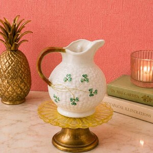 May include: A white ceramic pitcher with a textured basket-weave pattern and painted green shamrock motifs. The pitcher features a curved brown handle and sits atop a yellow glass pedestal stand with a brass-colored metal base.