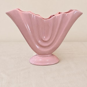 May include: A pale pink ceramic vase with a wide, fan-shaped opening and a scalloped, wavy rim. The body of the vase features vertical, curved ridges that extend from the center toward the edges, supported by a circular, pedestal-style base.