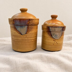 May include: Two cylindrical ceramic canisters with matching lids, featuring a warm honey-brown glaze with horizontal brushstroke textures and a distinct blue-grey drip glaze accent near the rim of each vessel, showcasing a handcrafted aesthetic.