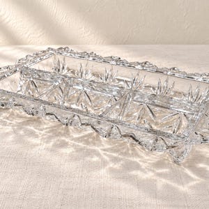 May include: A rectangular clear glass divided serving tray featuring a faceted starburst pattern on the base and a scalloped, serrated edge. The tray is partitioned into three distinct sections, showcasing a reflective, transparent crystalline texture.